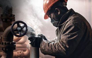 Worker in an orange hard hat, wearing a respirator mask and gloves, adjusting a valve on a pipe in an industrial setting with steam or mist in the background.